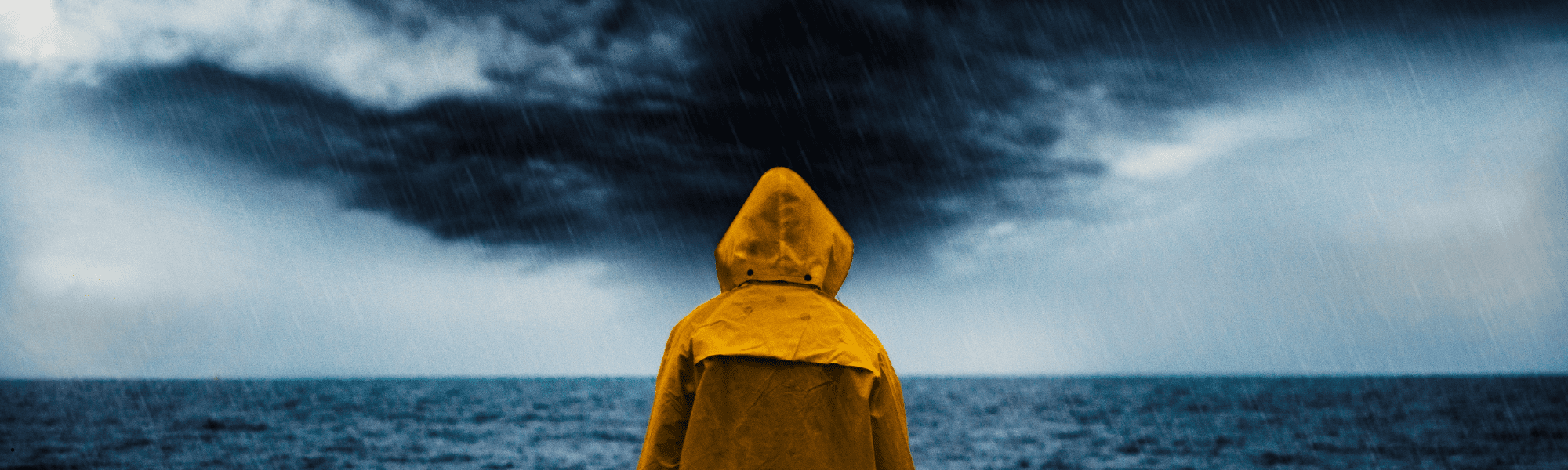Person in a yellow raincoat facing a storm over the ocean, rain falling heavily.